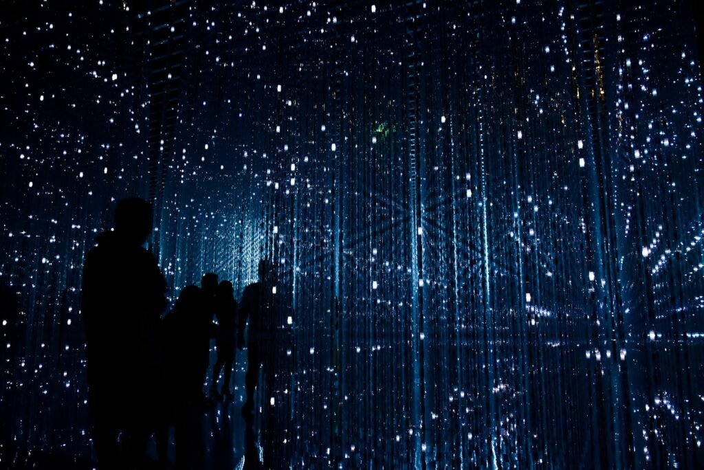 Silhouety lidí procházejících tmavým, futuristickým prostorem osvětleným tisíci drobných modrých světel, připomínajících hvězdnou oblohu.
