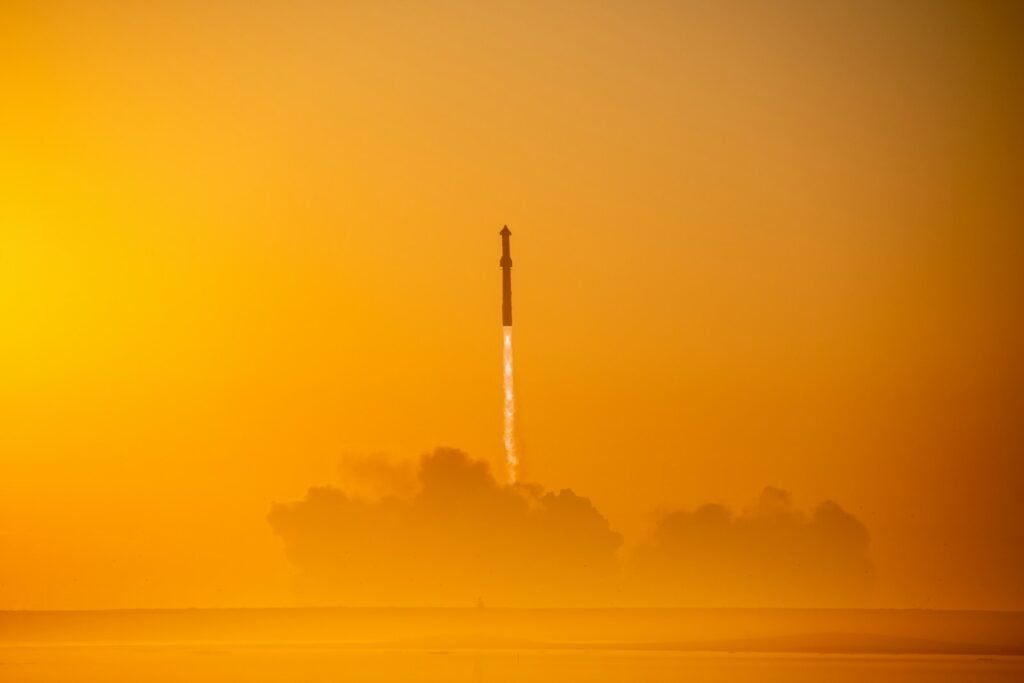 Start rakety na pozadí oranžové oblohy při východu nebo západu slunce, s oblakem kouře pod ní.